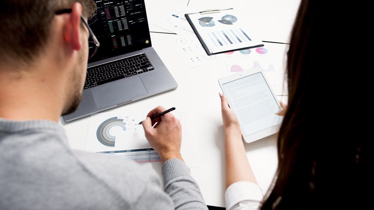 Services Two business professionals analyzing financial reports using a laptop and tablet in a modern office setting.
