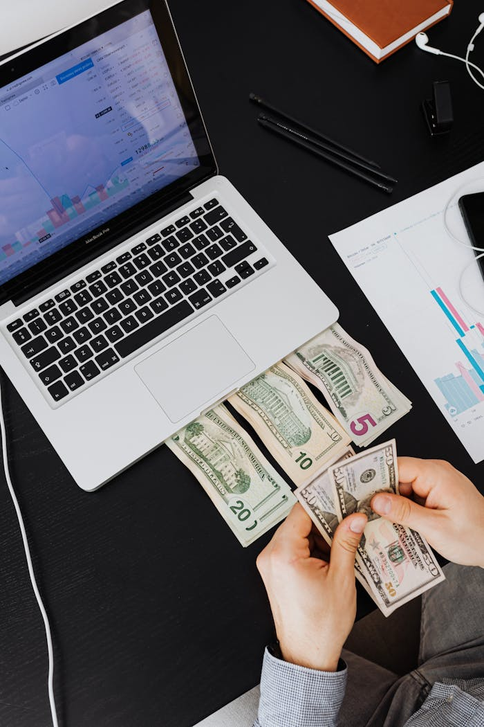 Home Person counting cash with laptop on desk, financial graphs visible.